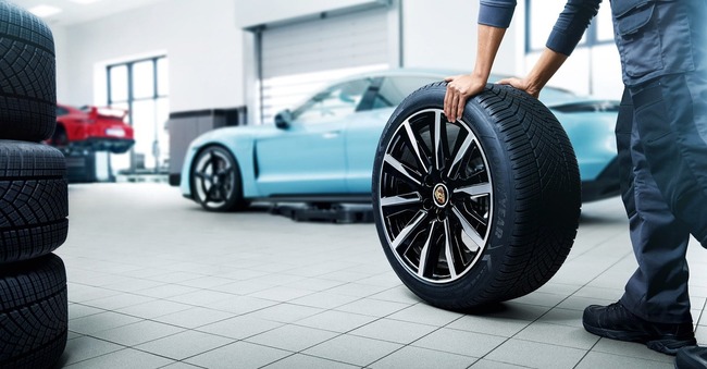 Porsche Certified Service Technician wheeling a Porsche tire toward a Tiffany blue Porsche vehicle for a tire change